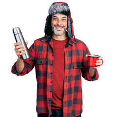 Wall Mural - Middle age hispanic man wearing hat with ear flaps drinking a cup of coffee holding thermo smiling with a happy and cool smile on face. showing teeth.