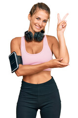 Wall Mural - Young blonde woman wearing gym clothes and using headphones smiling with happy face winking at the camera doing victory sign. number two.