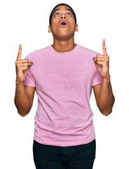 Wall Mural - Young handsome hispanic man wearing casual pink t shirt amazed and surprised looking up and pointing with fingers and raised arms.