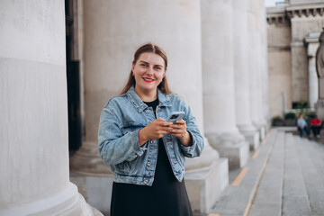 Wall Mural - Positive cheerful woman dressed in casual look holding smartphone gadget for communicate on the city street. Woman holding on cell smartphone while relaxing outdoors, Urban lifestyle concept