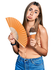 Canvas Print - Hispanic young woman holding hand fan eating ice cream depressed and worry for distress, crying angry and afraid. sad expression.