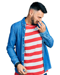 Wall Mural - Young hispanic man with beard wearing casual denim jacket smelling something stinky and disgusting, intolerable smell, holding breath with fingers on nose. bad smell