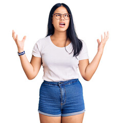 Wall Mural - Young beautiful asian girl wearing casual clothes and glasses crazy and mad shouting and yelling with aggressive expression and arms raised. frustration concept.