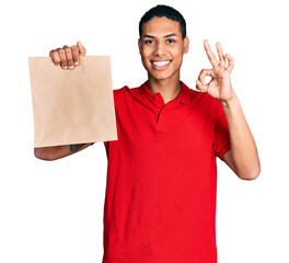 Young hispanic man holding take away paper bag doing ok sign with fingers, smiling friendly gesturing excellent symbol