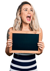 Canvas Print - Young blonde woman holding blackboard angry and mad screaming frustrated and furious, shouting with anger looking up.