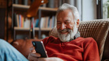 Fototapeta  - Close-up senior smiling relaxed retired man with beard sitting comfortably at home on armchair using mobile phone, communication concept