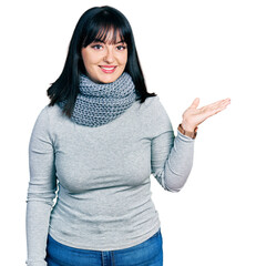 Poster - Young hispanic plus size woman wearing winter scarf smiling cheerful presenting and pointing with palm of hand looking at the camera.