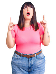 Wall Mural - Young plus size woman wearing casual clothes amazed and surprised looking up and pointing with fingers and raised arms.