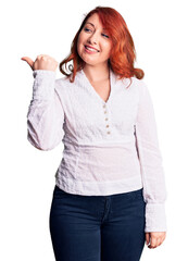 Canvas Print - Young beautiful redhead woman wearing casual shirt smiling with happy face looking and pointing to the side with thumb up.