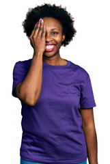 Wall Mural - African american woman with afro hair wearing casual purple t shirt covering one eye with hand, confident smile on face and surprise emotion.