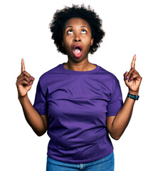 Poster - African american woman with afro hair wearing casual purple t shirt amazed and surprised looking up and pointing with fingers and raised arms.