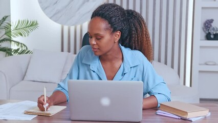 Wall Mural - Young successful casual African American business woman performs professional tasks using laptop with internet access and writing in diary sits at table in living room of apartment.