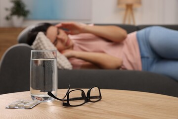 Canvas Print - Sad woman suffering from headache indoors, focus on glass of water, pills and eyeglasses