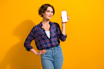 Wall Mural - Portrait of confident smart girl wear plaid shirt demonstrate smartphone touch screen empty space isolated on yellow color background