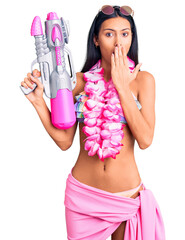 Wall Mural - Young beautiful latin girl wearing bikini and hawaiian lei holding water gun covering mouth with hand, shocked and afraid for mistake. surprised expression