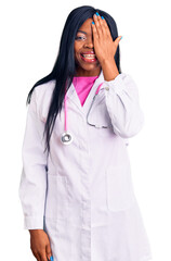 Wall Mural - Young african american woman wearing doctor stethoscope covering one eye with hand, confident smile on face and surprise emotion.