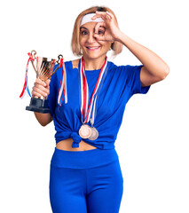 Poster - Young blonde woman holding champion trophy wearing medals smiling happy doing ok sign with hand on eye looking through fingers