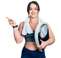 Wall Mural - Young hispanic girl wearing sportswear holding weighing machine smiling happy pointing with hand and finger to the side