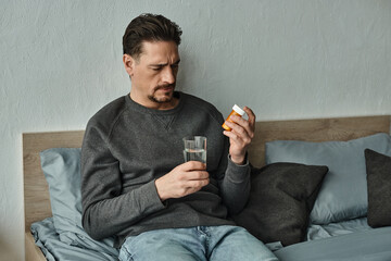 Wall Mural - bearded man in sweater holding glass of water and bottle with pills in bedroom, treatment