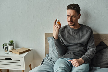 Wall Mural - pensive man looking at bottle with medication while sitting on bed in bedroom, treatment plan