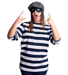 Sticker - Young beautiful brunette woman wearing burglar mask shouting with crazy expression doing rock symbol with hands up. music star. heavy concept.