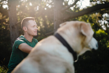 Wall Mural - Portrait of man with cute dog sitting in grass under tree. Happy owner of labrador retriever resting in nature on sunny summer day..