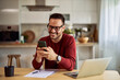 © Jelena - A smiling man sending text messages on his phone while working remotely over a laptop.