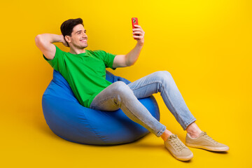 full length photo of cool man dressed green t-shirt jeans sit on pouf making selfie on smartphone is