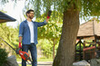 Smiling attractive man in gloves facing sun, while carrying garden equipment at backyard. Front view of dark haired joyful handyman with bush cutter holding hand up to block sun rays. Concept of work.