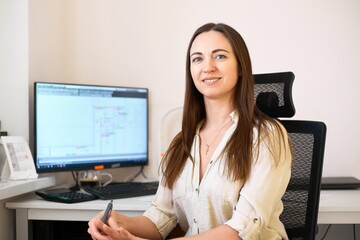 Wall Mural - Portrait of a remote worker. A beautiful woman sits at a table in front of a computer and designs a building. Confident young lady freelancer working from home.