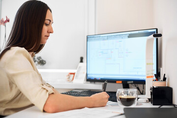 Wall Mural - Portrait of a remote worker. A beautiful woman sits at a table in front of a computer and designs a building. Confident young lady freelancer working from home.