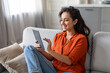 © Prostock-studio - Smiling woman in orange shirt using tablet on comfortable sofa at home
