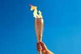 Fototapeta  - A hand holds the Olympic flame against a blue sky background, a torch in his hand, a symbol of the international sports games