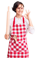 Wall Mural - Beautiful young woman with short hair wearing professional baker apron holding kneading roll doing ok sign with fingers, smiling friendly gesturing excellent symbol