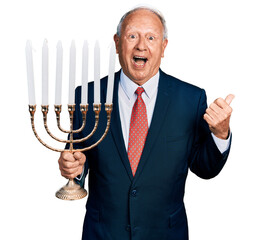 Poster - Senior business man with grey hair holding menorah hanukkah jewish candle pointing thumb up to the side smiling happy with open mouth