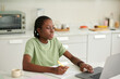 © DragonImages - Black teenage girl studying at home, reading article on laptop and writing in copybook