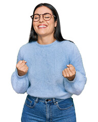 Wall Mural - Young hispanic woman wearing casual clothes and glasses very happy and excited doing winner gesture with arms raised, smiling and screaming for success. celebration concept.