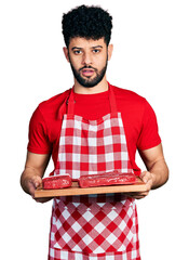 Poster - Young arab man with beard holding board with raw meat in shock face, looking skeptical and sarcastic, surprised with open mouth