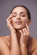 © Elliott/peopleimages.com - Woman, hands on face and beauty, skin with dermatology and natural cosmetics on purple background. Healthy glow, skincare with smile and wellness, self care and grooming for hygiene in studio