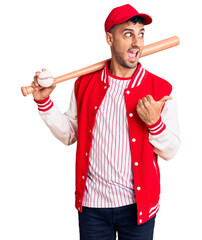 Wall Mural - Young hispanic man playing baseball holding bat and ball pointing thumb up to the side smiling happy with open mouth