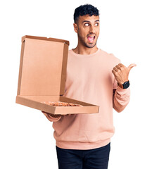 Wall Mural - Young hispanic man holding delivery pizza box pointing thumb up to the side smiling happy with open mouth