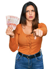 Sticker - Beautiful hispanic woman holding 10 colombian pesos banknotes pointing with finger to the camera and to you, confident gesture looking serious