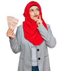 Poster - Beautiful hispanic woman wearing islamic hijab holding turkish lira banknotes serious face thinking about question with hand on chin, thoughtful about confusing idea