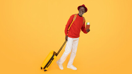 Wall Mural - Joyful senior man in a red hat and sweater, walking with a yellow suitcase and holding a passport