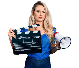 Poster - Young blonde woman holding video film clapboard and megaphone skeptic and nervous, frowning upset because of problem. negative person.