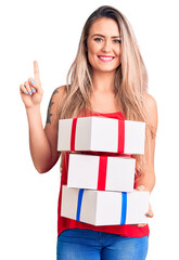 Poster - Young beautiful blonde woman holding birthday gifts surprised with an idea or question pointing finger with happy face, number one