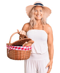 Wall Mural - Middle age fit blonde woman wearing summer hat and holding picnic wicker basket with bread looking positive and happy standing and smiling with a confident smile showing teeth