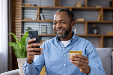 Wall Mural - A smiling African American man is at home, sitting on the sofa, holding a credit card and using a mobile phone. Close-up photo.