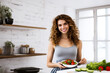 A charming smiling brunette is standing in the kitchen. A young girl prepares a salad of vegetables