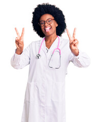 Wall Mural - Young african american woman wearing doctor coat and stethoscope smiling with tongue out showing fingers of both hands doing victory sign. number two.